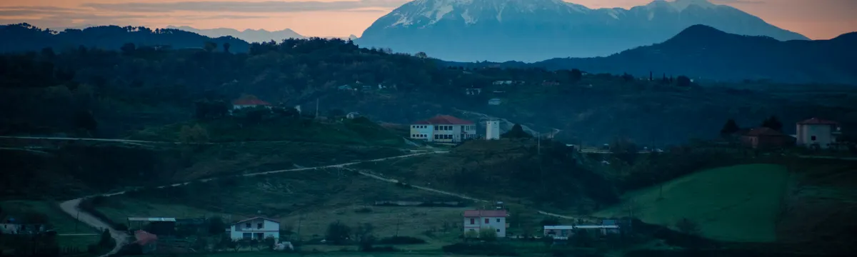 אלבניה: A tranquil rural scene in Albania, showcasing rolling hills and mountains under a sunset sky.