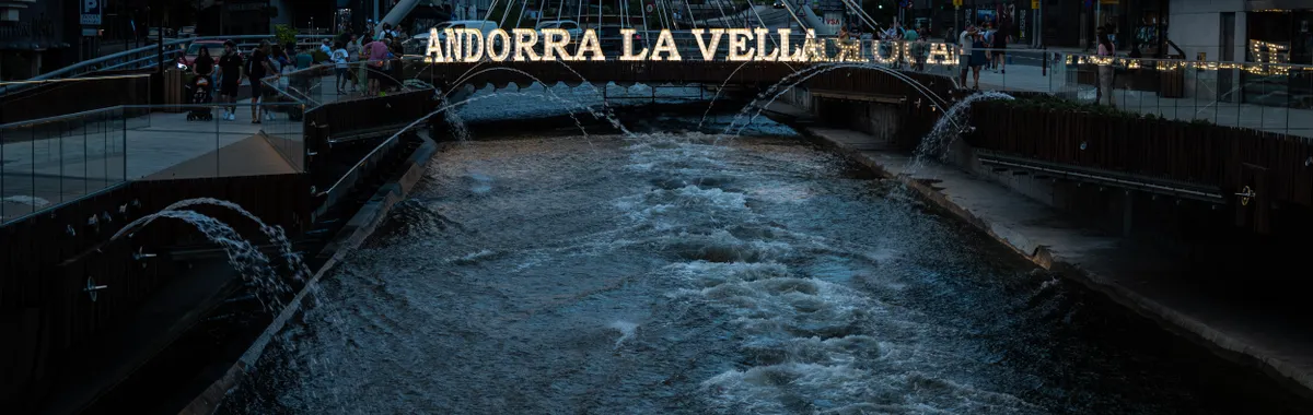 Night view of the iconic Andorra la Vella bridge over the urban river, with city lights and flowing water.