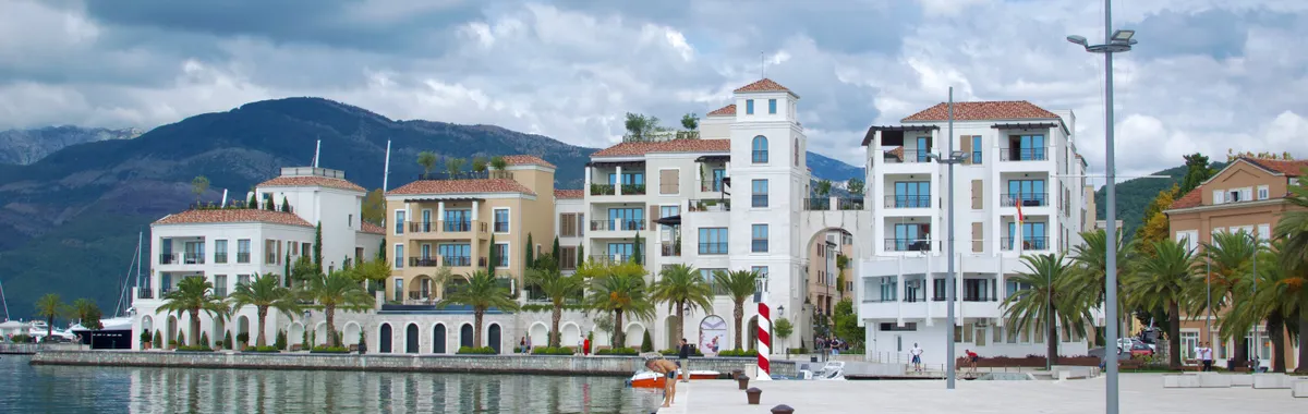 Beautiful waterfront buildings in Tivat, Montenegro, with mountains and a cloudy sky.