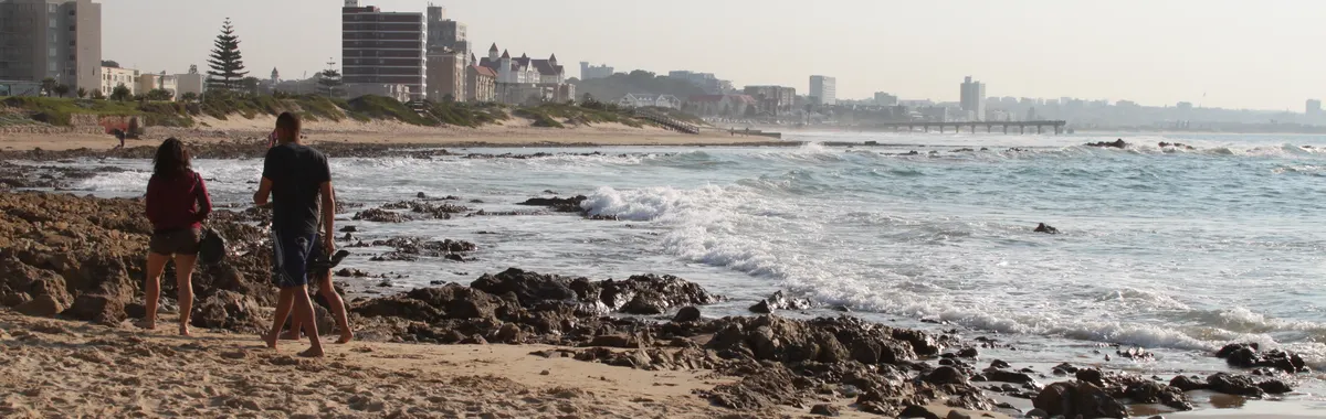 A scenic view of people walking along Gqeberha Beach with city skyline in the background.