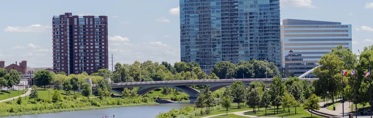Scenic view of a modern urban skyline by a riverside with lush greenery and blue sky.