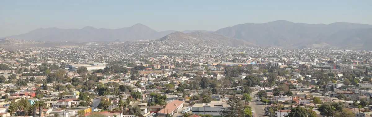 Aerial daytime view of Tijuana's cityscape with mountainous backdrop under a clear sky.
