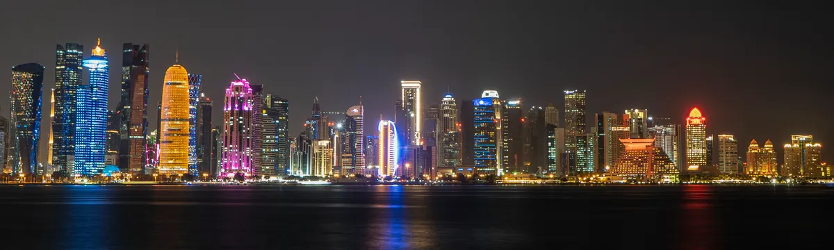 קטאר: Doha cityscape featuring illuminated skyscrapers reflecting on the water at night.